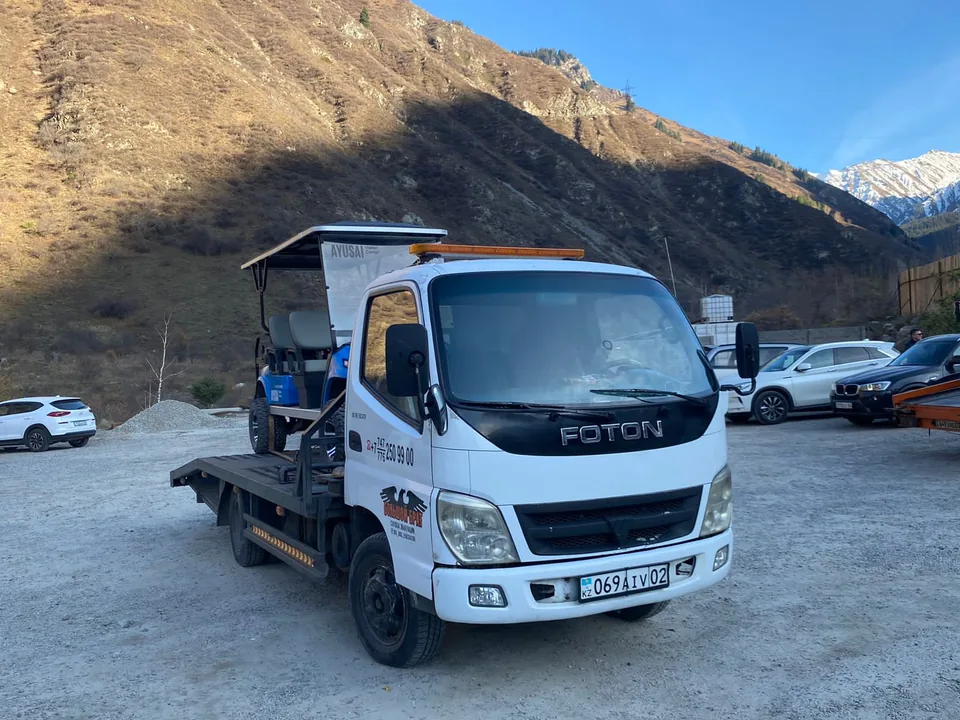Белый эвакуатор BIG BRO Foton с гольф-каром на платформе на фоне гор